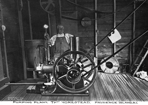 A mustachioed man in coveralls monitors a pressure gauge and the machinery at a pumping plant. Caption reads: "Pumping Plant. The Homestead. Prudence Island, R.I."