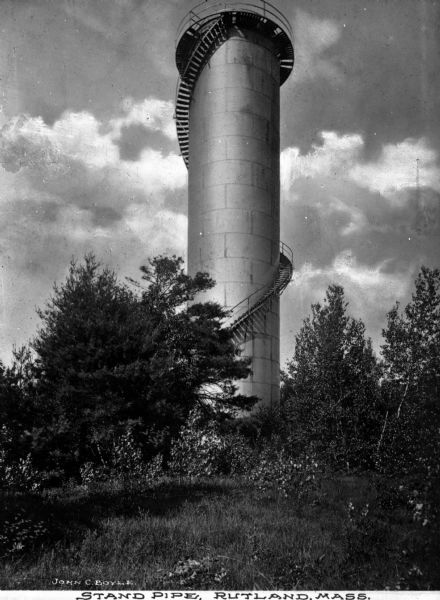 Stairs wrap around a tall cylindrical water tower in a grassy setting. Caption reads: "Stand Pipe, Rutland, Mass."