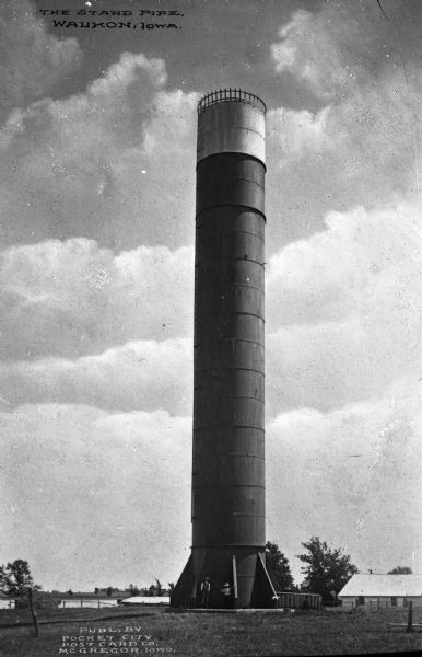 Two men stand at the base of a tall standpipe, also known as a water tower. Caption reads: "The Stand Pipe, Waukon, Iowa."