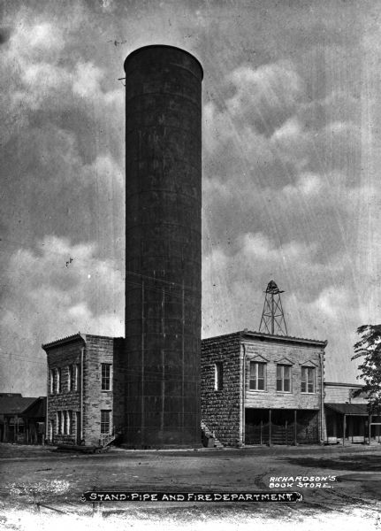 A tall standpipe, also known as a water tower, next to a fire department. Stables are at the street level and a bell is on the roof. Caption reads: "Stand Pipe and Fire Department."