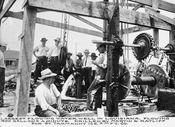 A group of men wearing hats gathered around a water pump. Scaffolding surrounds the operation and a large pulley is visible. Text on image reads "Largest flowing water well in Louisiana, flowing 400 gallons a minute drilled by Martin & Ratliff for St. Tammanny Ice & M.F.G. Co."