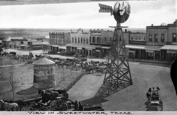 Elevated view of a small town area with a windmill in the main square lined with store fronts including a drug/book store and a hardware/lumber store. A small silo is visible and is emblazoned with the words "Burtonling (obscured) Lumber." Horses and horse-carts are gathered throughout the square. Caption reads: "View in Sweetwater, Texas."