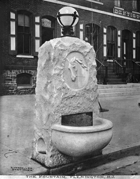 View of a stone fountain with a light fixture, presumably for horses, on a curb in front of a "Clerk's Office" and a "Dentist." A horse's head is carved into the stone. Caption reads: "The Fountain, Flemington, N.J."