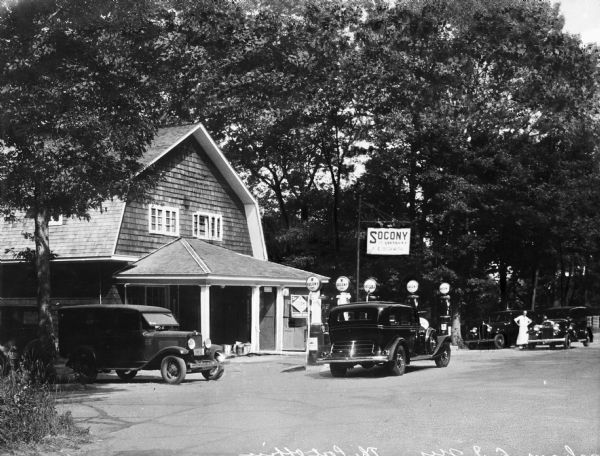 Exterior of a shingled post office/gas station with cars parked in front near the gas pumps. Sign in front reads "Socony" and a man wearing a white apron and hat is standing near a car on the far right.