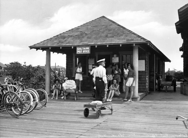 A postal worker using a wagon to bring letters to the small Saltaire Post Office building. People are standing and sitting on the porch area and bikes are visible to the left side of the image.
