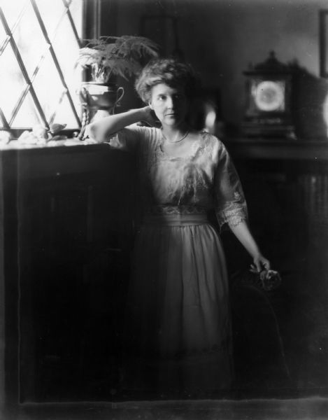 Three-quarter length portrait of Zona Gale (Mrs. William Breese) standing near a window and holding a rose in her left hand.