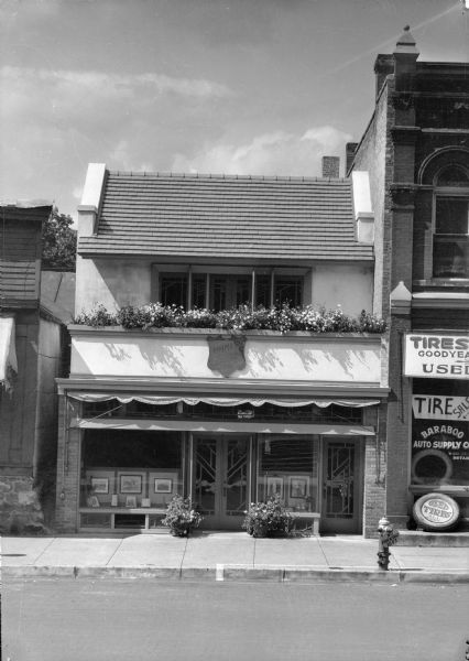 A view of the exterior of the Trimpey photographic studio and antique shop, 128 Fourth Street. The second story is set back, with a porch and flower boxes in front. A distinct Prairie Style influence is seen in the doors and windows. Examples of Trimpey's photography are displayed in the front windows. Baraboo Auto Supply Company occupies the building on the right.