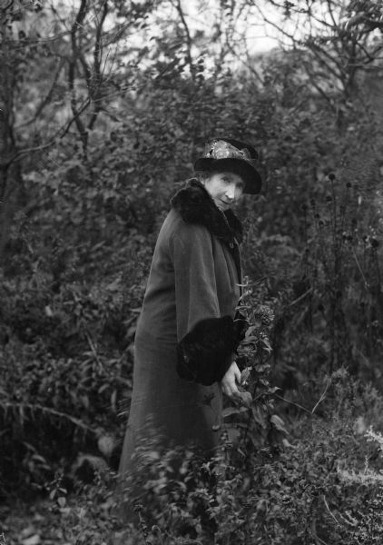 Mrs. Ephraim Burt Trimpey points to a zinnia blooming in a late summer garden. She us wearing a hat and fur-trimmed coat.