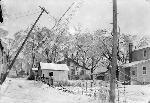 Winter scene of a Baraboo, Wisconsin, alley after an ice storm, possibly March 2, 1922. Utility poles are leaning and their wires sag under the weight of the ice. Houses, sheds, and fences show no damage from the storm.