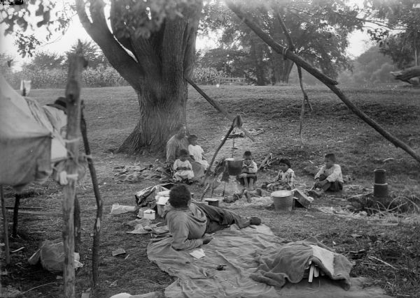 A Native American (possibly Ho-Chunk) family rests at their campsite under a large tree. There is a bucket suspended over a small campfire.