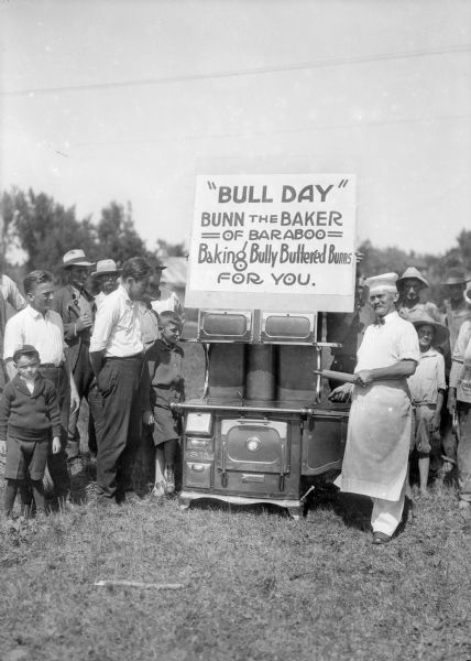 Bunn the Baker, in chef's hat and apron and holding a rolling pin, stands beside a wood burning cook stove set up outdoors. A sign above the stove advertises "Bully Buttered Bunns," and a crowd of men and boys stands by. John Bunn operated a bakery at 114 Walnut Street in Baraboo.