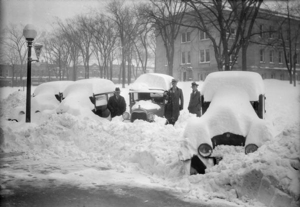 Winter scene with three men standing beside parked cars that have been buried by snowfall and snowplow on Fourth Avenue, Baraboo, Wisconsin. The Sauk County Courthouse is on the right; commercial buildings along Oak Street may be seen in the background. A sign on a lamppost identifies Highway 33 West.