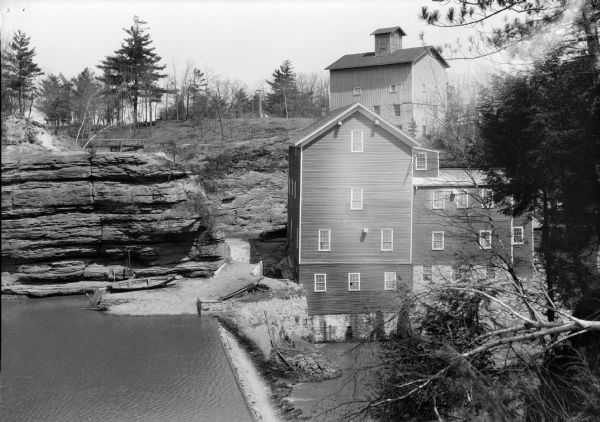Elevated view of the Timme Mill and dam on Dell Creek at Mirror Lake.  There is a large building with cupola, probably a granary, on the hill behind the mill. A boat rests at the foot of a rock outcropping.