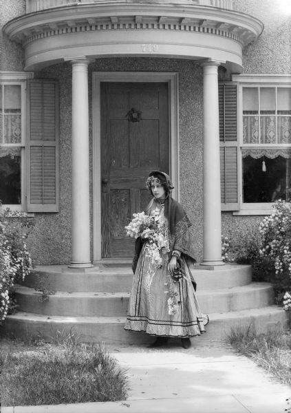 An unidentified young woman in old-fashioned dress holds flowers as she stands in front of entrance of the Trimpey house at 719 Oak Street.