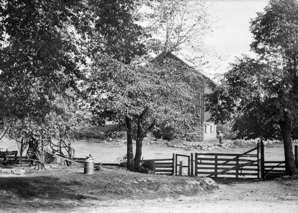 A farm scene with a fence and gate, and milk can in the foreground. In the background is a large barn with a stone foundation, with a man walking nearby.