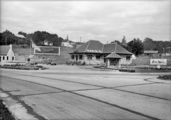 View from across road of the Devil's Barbecue roadside restaurant on South Boulevard, then Highway 12, near Baraboo. The restaurant is a stone building; there is a small stand selling Hires Root Beer and Coca-Cola across the driveway. A car is parked in the background near a billboard advertises a garage.