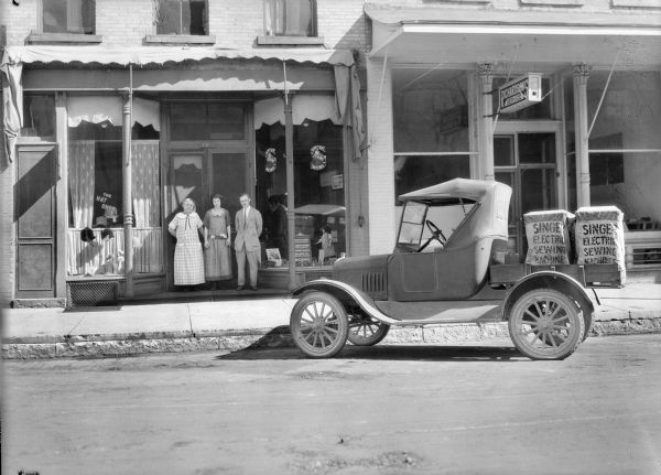 View from across street of a small truck loaded with two "Singer Electric Sewing Machines" parked in front of a store.  The store, identified as "The Hat Shop," advertises Singer Sewing Machine accessories and repairs. A man and two women pose in the doorway.