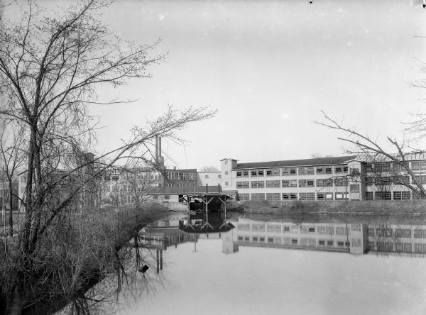 View across river of a large mill or factory complex, possibly the Island Woolen Mill. In front of the mill is a roofed boathouse, with a canoe hanging from the ceiling.