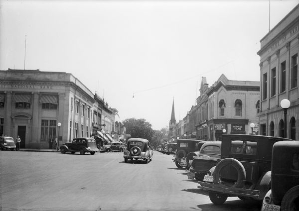 View down street of cars and trucks parked along Third Street in this view looking East across Oak Street. The First National Bank, left, is diagonally across the intersection from the Bank of Baraboo. The Corner Drug Store, on the southeast corner, advertises Kodak film and sodas.