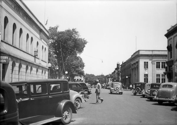 A man crosses Oak Street in this view north across the intersection with Third Street. The Bank of Baraboo is on the left; First National Bank is diagonally across the intersection. Trees on the Sauk County Courthouse square are on the left. Cars fill the angle parking spaces on both sides of the street.