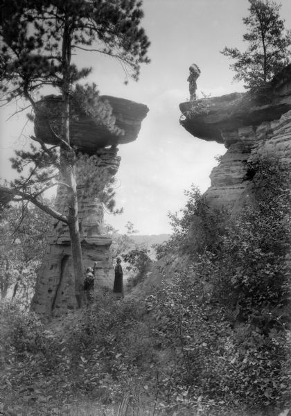 A man in Native American clothing is standing on the ledge near Stand Rock. Two women are watching from the ground below.