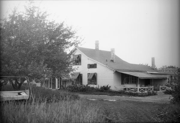The rear of Helen Baldwin's Restaurant near Baraboo. Notes on the negative envelope indicate that the restaurant was operated by the photographer and his wife, Alice Trimpey. There is a lawn swing in the yard and also on the awning-roofed porch. Flowers bloom along the wall of the restaurant.