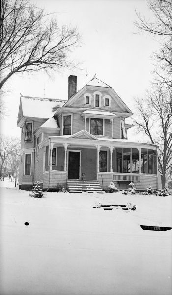 View from street of a dog sitting on the front porch of a large Victorian house at 127 Eleventh Street, the home of Dr. Clausen F. Stekl, a Baraboo dentist. A screened porch curves around the right side of the house. There is decorative shingling in the front gable.