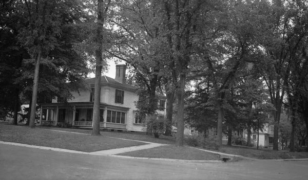 View from street of a two-story, wood frame house with a front porch sittomg on a wooded lot. The house has a large chimney, dentil molding under the eaves, and Greek Revival style corner pilasters. Bushes have been planted in front of the entry onto the porch.