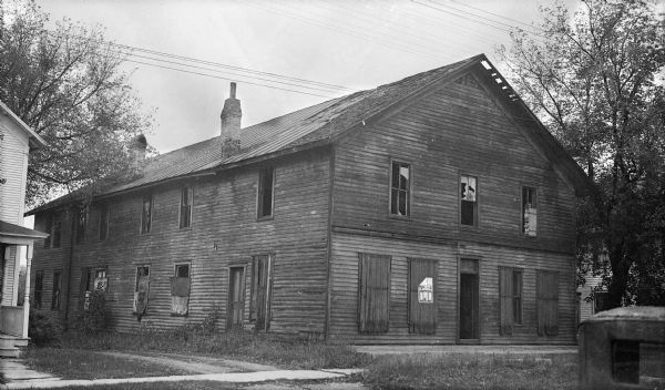 A deteriorating, large, two-story building with Greek revival features, which may have served as an inn or hotel. The windows are boarded up or broken and the roof is failing. An advertisement has been posted on the front.