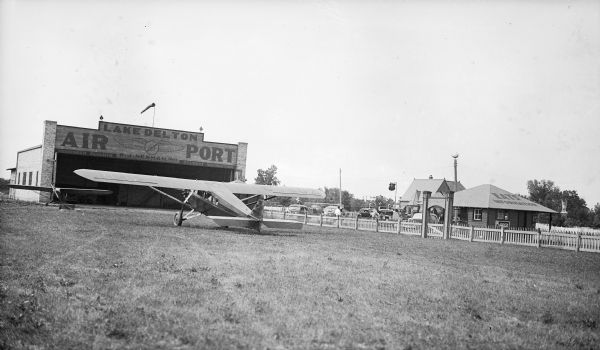 View across field towards two airplanes sitting in front of the hangar at the Lake Delton Airport. The sign on the hangar identifies W.J. Newman as the owner, and advertises passenger flights and student instruction. Cars are parked outside of the fence on the right. A small building advertises "Eats" and "Information" on its roof; a Coca-Cola machine is on the porch.