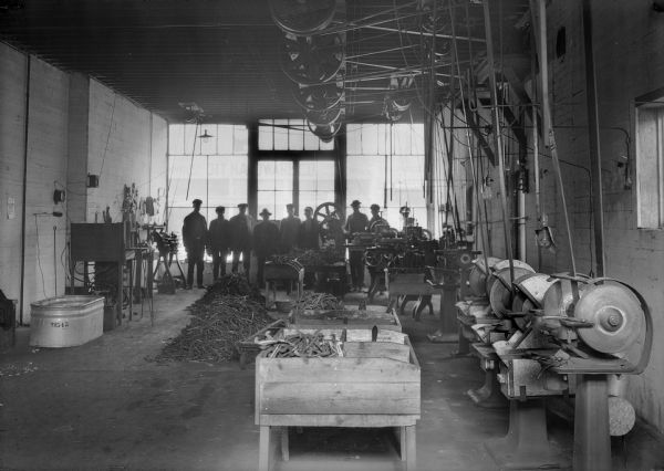 A group of eight men pose in the back of a room against a row of windows in a machine shop. There are overhead belts that power a row of grind stones; metal parts, possibly castings, fill wooden bins and are stacked on the floor. The building opposite has a sign which reads "Ott Hardware Co."