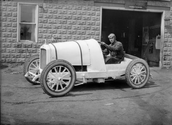 A man in racing attire and goggles sits behind the wheel of a race car. There is a cement block building behind him with an open garage door.
