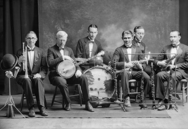 Six musicians pose in front of a backdrop with their instruments, including saxophones, clarinets, a banjo, drums and trumpet with mutes.  The drum has been painted with a seascape and the word "Orchestra" in faux Oriental letters.