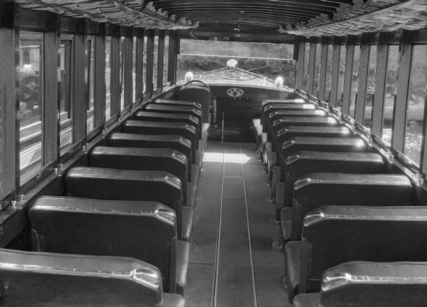 The interior of an enclosed tourist motor launch, possibly the <i>Seagull</i>. Coach type seats line the sides. A glimpse of water and shoreline is visable through the windows on the right.