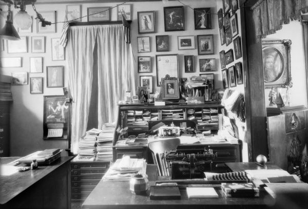 An interior view of a cluttered but organized office, possibly that of the photographer. There are prints of female nudes and bathing beauties on the far wall. A curtained doorway is adorned with two American flags. There is an open roll top desk and also a typewriter desk with a Remington typewriter on it. An extension cord has been attached to a socket in the overhead light fixture.