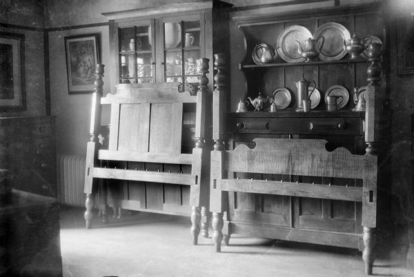 The head and foot boards of a maple bed lean against two cupboards.  The cupboards hold pewter ware as well as crockery.