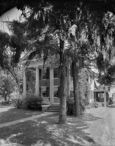 A stately house in the Greek Revival style stands under large evergreen trees. There are shutters on the windows and a two-story, columned front porch.