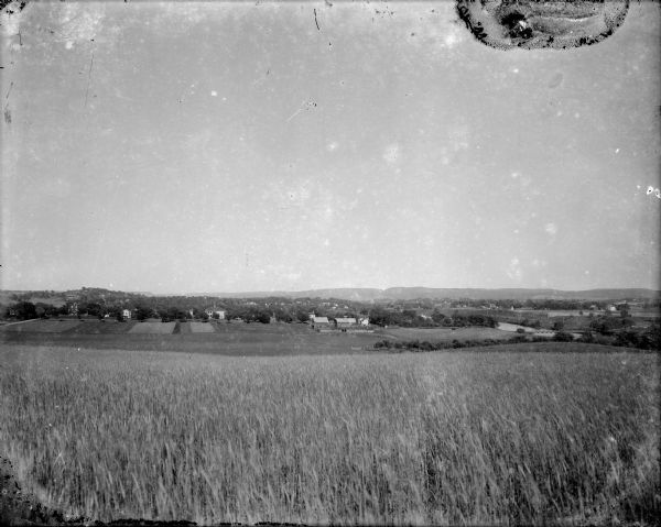 A view of a rolling rural landscape, most likely in Sauk County. In the far distance are houses and farm buildings, a church and a larger building, center left, possibly a school. On the right, there is a bridge over a small river. In the foreground is a grain field, and there are taller hills or bluffs in the background.