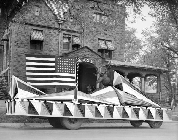 A parade float decorated with bunting, a large, 48-star American flag, and a mounted elk's head is parked in front of the Baraboo Elks Lodge.  The large stone house was originally the home of Al. Ringling.  "B.P.O.E." above the entrance identifies the Benevolent and Protective Order of Elks. Two men sit on the front porch.