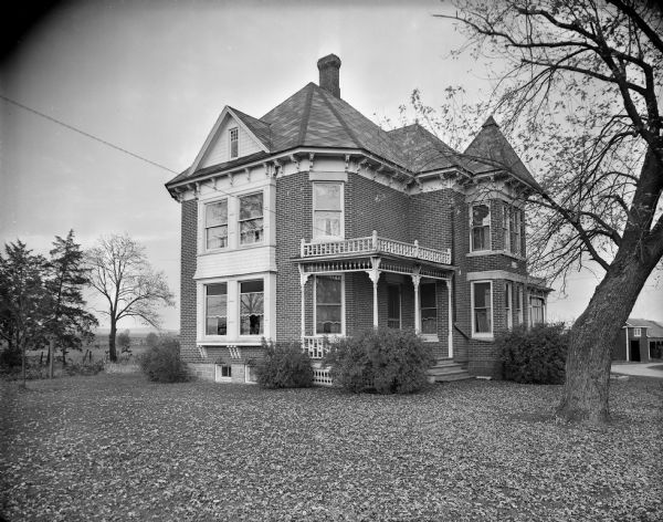A two-story Victorian brick farmhouse with eave brackets, bay windows and a front porch with fretwork railing. The Baraboo bluffs are visible in the distance.