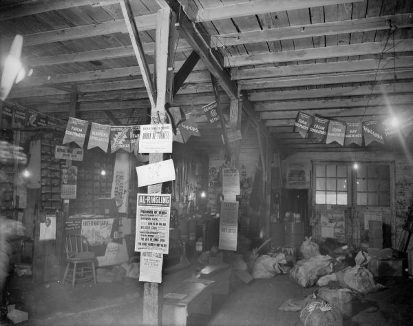 An interior view of an unidentified farm supply store and International dealership, with banners advertising McCormick-Deering farm implements and tractors. There are various bills posted for upcoming attractions at the Al. Ringling Theatre, auctions, etc.