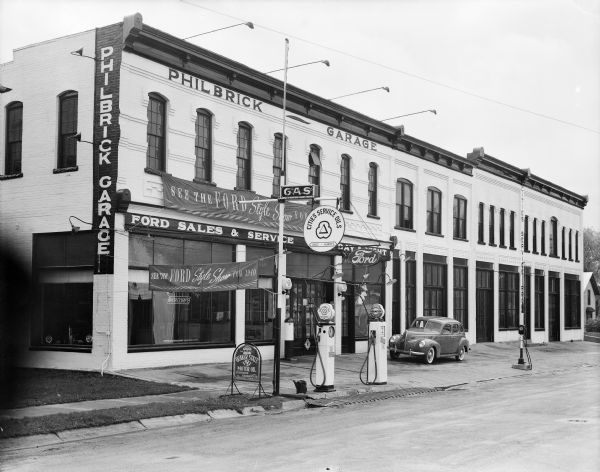 An exterior view of the Philbrick Garage, a Ford dealership and Cities Service filling station located at 207 Third Avenue. A sign advertises Quaker State Motor Oil. A new Ford automobile with whitewall tires is parked in front of the building. The business occupies four attached, two-story brick storefront buildings.