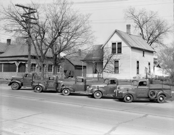 A fleet of four pickup trucks of various makes and a larger truck, all advertising L.C. Welch Plumbing and Heating on the door. They are parked on a residential street.