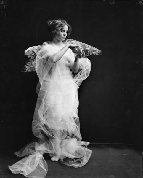 A young woman stands holding a sea shell. She wears wings and is loosely wrapped in diaphanous cloth.