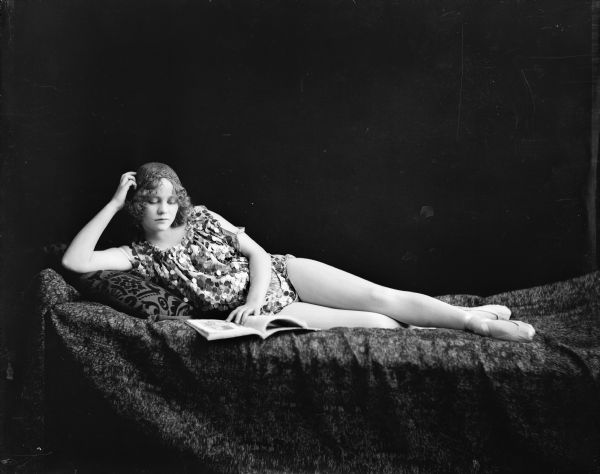 A young woman poses reclining and reading a magazine. She wears a bangled costume, tights, and dance slippers.