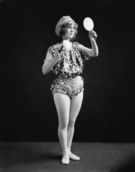 A woman poses with a mirror and powder puff. She wears a bangled costume and dance slippers.