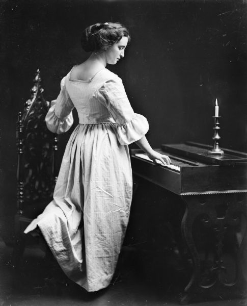 A woman in an old style dress stands in profile by a keyboard instrument, with her left knee resting on a Gothic Revival chair. A candle is burning in a brass candlestick on the instrument.