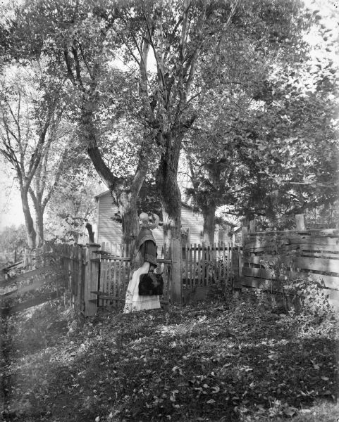 A woman in old style dress with bonnet, cape and carpet bag, stands at a gate in a picket fence. There is an old house in the background.