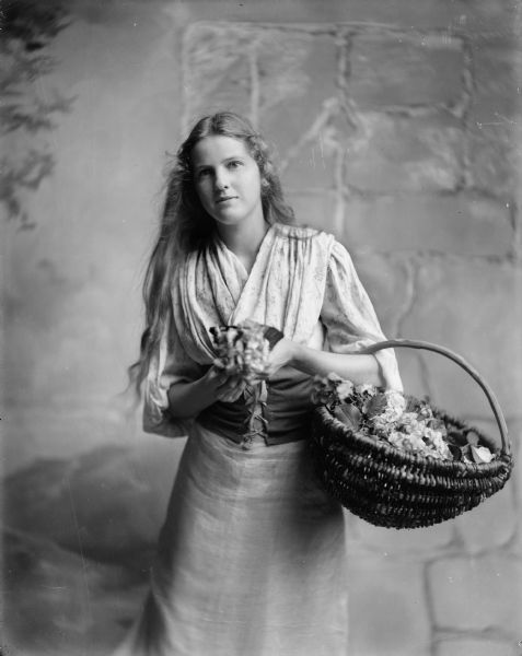 A young woman poses with a basket of flowers over one arm. She wears an old-fashioned costume and stands in front of a painted backdrop which mimics a stone wall.