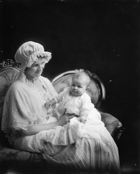 A young woman in night cap and dressing gown holds an infant in a long white dress. She is seated on a Victorian settee.